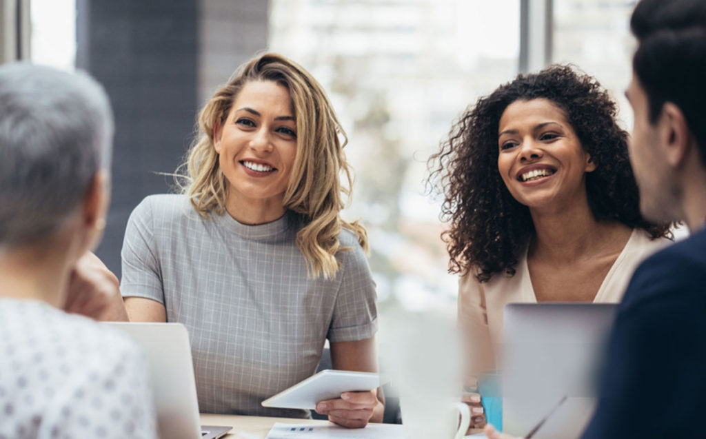 A group of four people are sitting at a table in an office environment, engaging in a conversation and smiling. Two women are holding electronic devices, possibly discussing a business broker franchise opportunity.