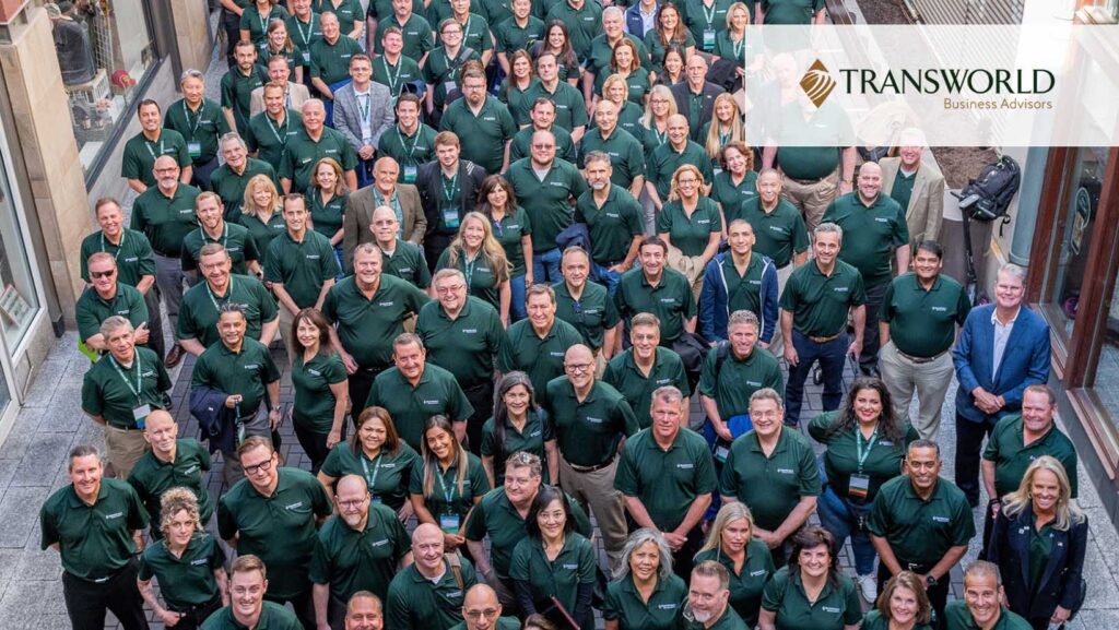 A large group of people in green shirts stands closely together, smiling, in an outdoor setting, embodying the spirit of a dedicated business broker team. A "Transworld Business Advisors" logo is visible in the top right corner.