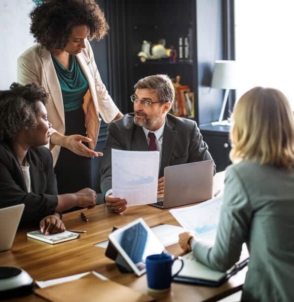 Four professionals in business attire are having a meeting around a table with laptops and documents. One man holds a paper, and a woman stands, gesturing towards it.