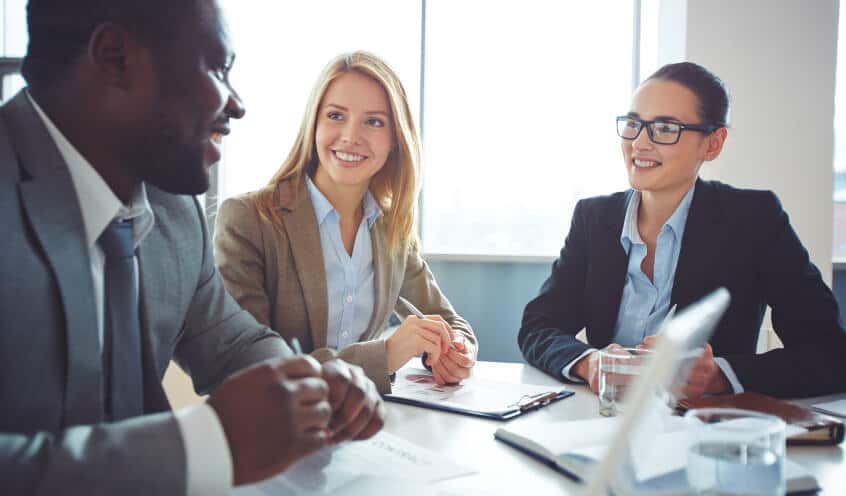 Three professionals are sitting around a table in a meeting room, engaged in a discussion. Two women are visible, one smiling, and a man is speaking. Papers and a glass of water are on the table.