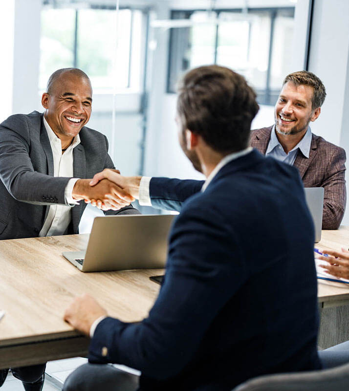 Two men smile and shake hands across a table in an office setting, with a third man seated beside them, observing.