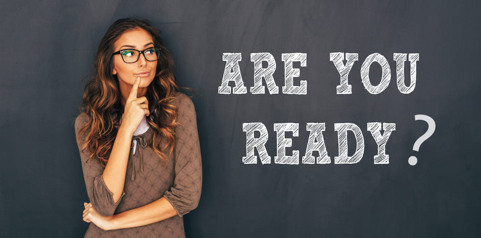 Woman contemplating with her finger on her chin in front of a chalkboard with the text "ARE YOU READY?.