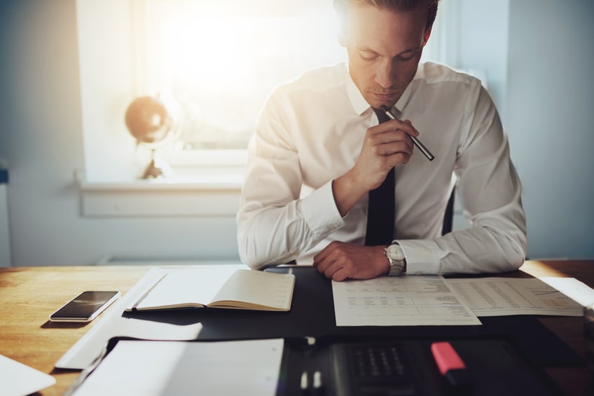 A man in a shirt and tie sits at a desk reviewing documents, with a notebook, phone, and stationery nearby, sunlight streaming through a window.