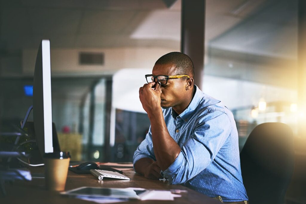 A person sitting at a desk, rubbing their eyes while looking at a computer monitor, with a coffee cup beside them. The setting appears to be an office.