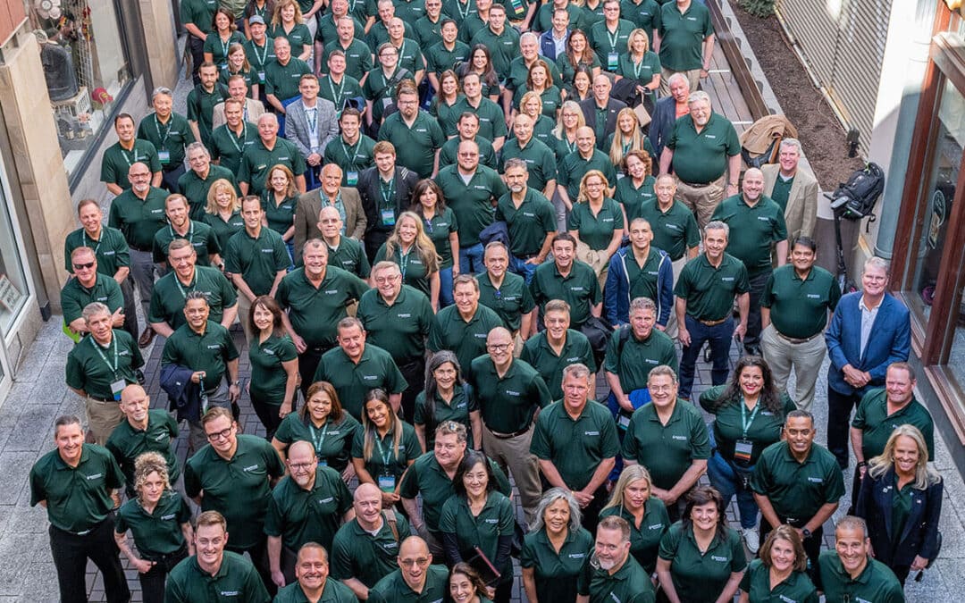 A large group of people wearing matching green shirts stands closely together outdoors for a group photo, looking up at the camera.